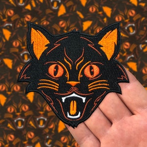 Könnte beinhalten: Schwarzer gestickter Bügeleisen-Aufnäher mit einem schwarzen Kater mit orangefarbenen Augen und Mund sowie orangefarbenen Ohren und Schnurrhaaren. Die Katze grinst mit herausgestreckter Zunge.