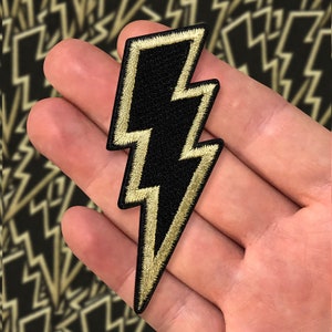 May include: A black and gold embroidered lightning bolt patch. The patch has a black centre with gold-coloured stitching around the edges. The background shows a pattern of lightning bolts.