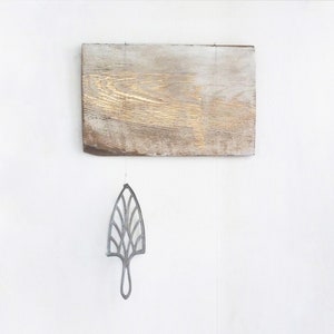 May include: A silver metal leaf-shaped object hangs from a string attached to a wooden board. A black metal weight hangs from a string below the leaf-shaped object.