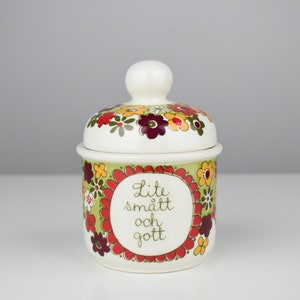 以下が含まれることがあります： 花柄の白いセラミック製の壺と蓋。壺は緑色の背景に赤、オレンジ、黄色の花が描かれている。蓋は白いノブと同様の花柄。壺には「Lite smalt och gott」という文字が書かれている。