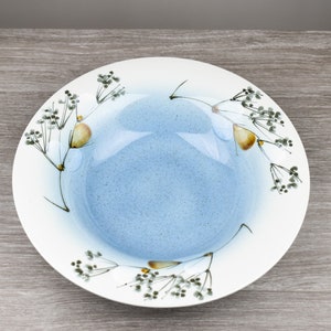Può includere: Una ciotola in ceramica bianca con una glassa blu e un motivo floreale. La ciotola ha un bordo largo e una profondità poco profonda. Il motivo floreale presenta rami delicati con piccoli fiori gialli e foglie verdi.