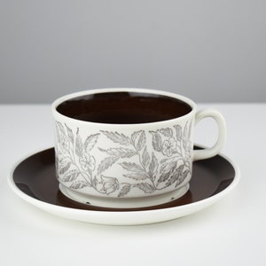 May include: A white teacup and saucer with a brown rim and a floral pattern in grey. The cup has a brown interior.