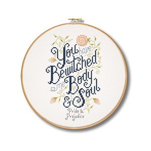 May include: A white embroidery hoop with a navy blue cross-stitch design that reads "You have bewitched me body & soul" with floral accents. The text "Pride & Prejudice" is written below the design.