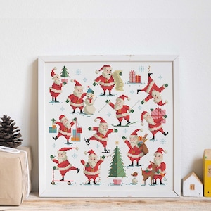 May include: A white frame containing a cross-stitch design of Santa Claus in various poses. The design includes a snowman, a Christmas tree, and presents. The frame is on a wooden surface with a brown gift box, a pine cone, and small wooden houses.