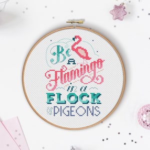May include: A white embroidery hoop with a cross-stitch design that reads "Be a flamingo in a flock of pigeons". The design features a pink flamingo, blue and green lettering, and a white background.