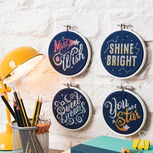 Puede incluir: Cuatro aros de bordado redondos con tela azul y letras blancas. Los aros están colgados en una pared de ladrillo blanco. Las letras dicen "Make a Wish", "Shine Bright", "Sweet Dreams" y "You are a Star".
