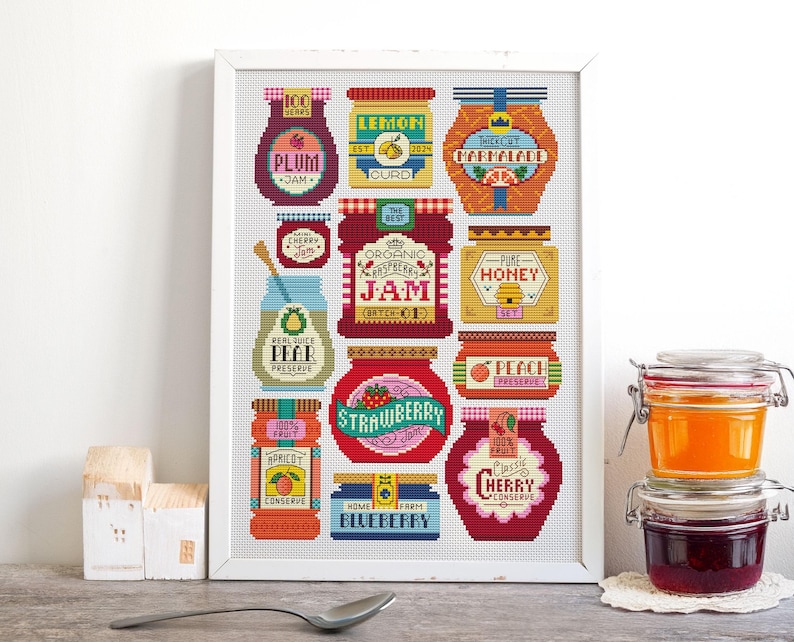 May include: A framed cross-stitch artwork featuring colorful jars of preserves, including plum jam, lemon curd, and strawberry jam. The artwork is in a white frame and includes text such as "Organic Raspberry Jam" and "Pure Honey". Jars of preserves are stacked on a white doily.