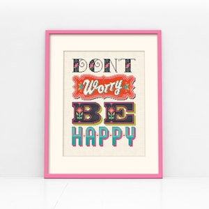 May include: A framed cross-stitch design with a pink frame. The cross-stitch reads "Don't Worry Be Happy" in colorful, stylized lettering.