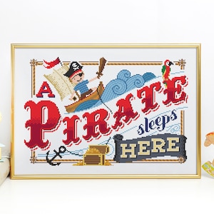 May include: Cross-stitch artwork in a gold frame. The design features a pirate in a boat, the words "A Pirate sleeps HERE", a treasure chest, and a parrot. The text and pirate are in red, blue, and gold.