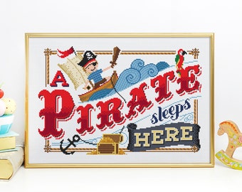 Könnte beinhalten: Kreuzstich-Kunstwerk in einem goldenen Rahmen. Das Design zeigt einen Piraten in einem Boot, die Worte "A Pirate sleeps HERE", eine Schatzkiste und einen Papagei. Der Text und der Pirat sind in Rot, Blau und Gold gehalten.