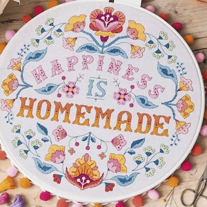 Pode incluir: Um aro de bordado branco com um design floral colorido e o texto "Happiness is Homemade".