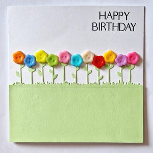 May include: A birthday card with the words "HAPPY BIRTHDAY" in black. The card features a row of colourful button flowers with green stems and leaves, set against a white background. A light green rectangle is at the bottom.