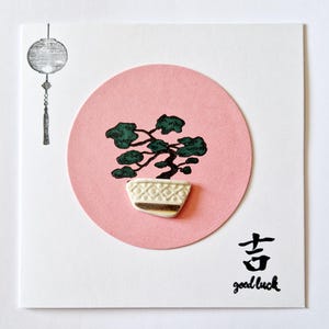 May include: A pink card featuring a green bonsai tree in a white pot. The card includes a Chinese character for "good luck" and the words "good luck" written beneath it.