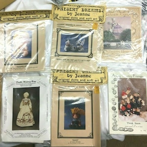 Może przedstawiać: Kolekcja ośmiu wzorów na lalki i miękką sztukę. Wzory znajdują się w plastikowych torebkach i przedstawiają różne projekty, w tym królika, lalkę z obrączką ślubną, lalkę w czerwonym kapeluszu i bałwana. Wzory są zatytułowane "Present Dreams by Jeanne", "Karen's Coop", "Present Dreams by Jeanne", "Huck" i "Among Country Friends".
