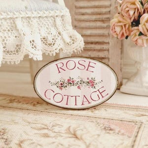 May include: An oval sign reading "ROSE COTTAGE" in pink, with a floral design. It sits on a floral rug, with a white vase of pink roses and a lace-trimmed chair behind it. The sign has a brown border. The scene evokes a cottage theme.