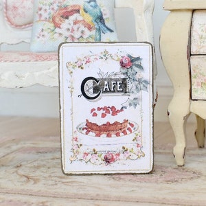May include: A vintage-style sign with a floral design and the word "Cafe" in a decorative font. The sign features a picture of a cake with strawberries on top.