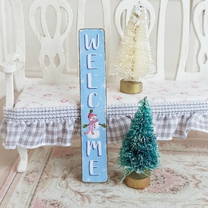 Puede incluir: Un letrero de madera azul en miniatura con la palabra "WELCOME" pintada en letras blancas. Un mu&ntilde;eco de nieve con un sombrero rojo y una bufanda est&aacute; pintado en el letrero. El letrero est&aacute; sentado en un banco blanco con un coj&iacute;n de estampado floral rosa y blanco. Hay dos &aacute;rboles de Navidad en miniatura en el banco, uno blanco y otro verde.