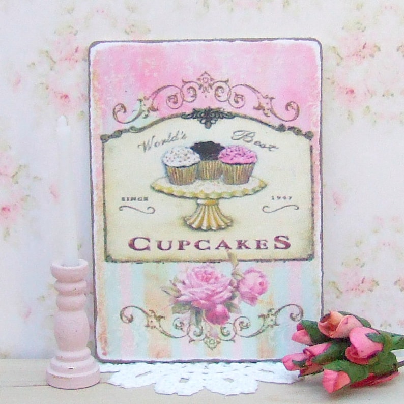 K&ouml;nnte beinhalten: Ein rechteckiges Schild mit rosa und hellblauem Hintergrund zeigt ein Cupcake-Design. Auf dem Schild steht "World's Best Cupcakes Since 1947". Ein rosa Kerzenhalter und ein Strau&szlig; rosa Rosen befinden sich im Vordergrund.