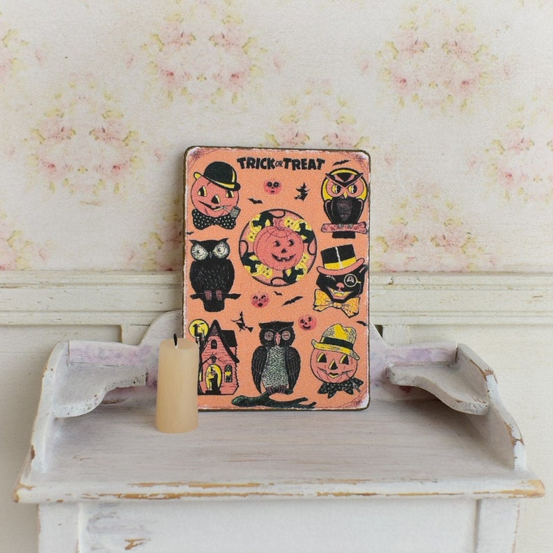 May include: A vintage-style Halloween sign with orange background and various spooky illustrations, including owls, pumpkins, and a black cat. The sign reads "TRICK OR TREAT" at the top. A cream-colored candle sits in front of the sign.