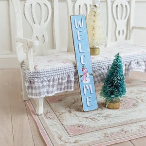 Puede incluir: Un banco blanco en miniatura con un letrero de bienvenida azul y blanco, un &aacute;rbol de Navidad blanco flocado y un &aacute;rbol de Navidad verde flocado. El banco tiene un coj&iacute;n a cuadros gris y blanco.