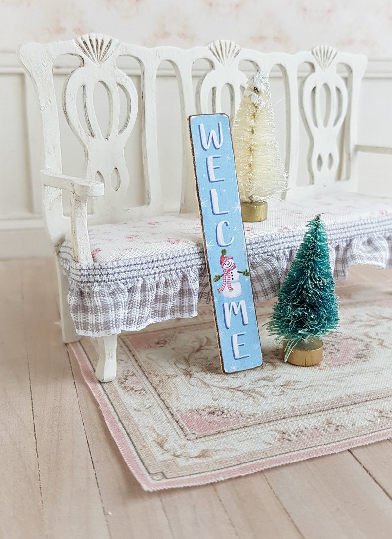 Puede incluir: Un banco blanco en miniatura con un letrero de bienvenida azul y blanco, un &aacute;rbol de Navidad blanco flocado y un &aacute;rbol de Navidad verde flocado. El banco tiene un coj&iacute;n a cuadros gris y blanco.