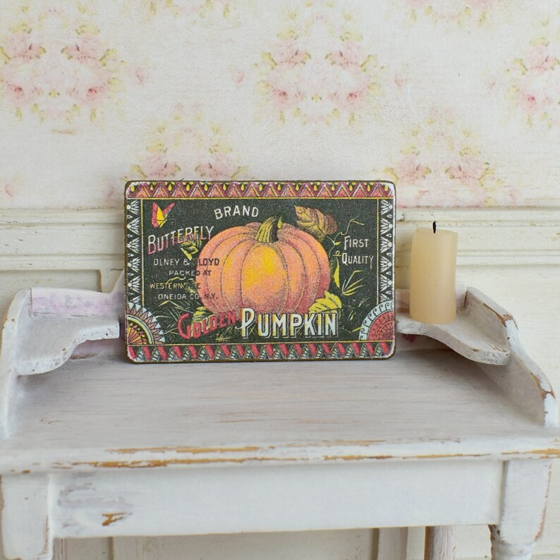 Puede incluir: Un letrero de estilo vintage con fondo negro y borde de colores. El letrero presenta una gran calabaza naranja con un tallo y hojas verdes. El texto en el letrero dice "Butterfly Brand Golden Pumpkin First Quality".