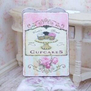 K&ouml;nnte beinhalten: Ein rechteckiges Schild mit rosa und hellblauem Hintergrund, mit dem Text "World's Best Cupcakes" und einer Illustration von drei Cupcakes auf einem Podest. Das Schild enth&auml;lt auch ein Blumendesign und die Worte "Since 1947".