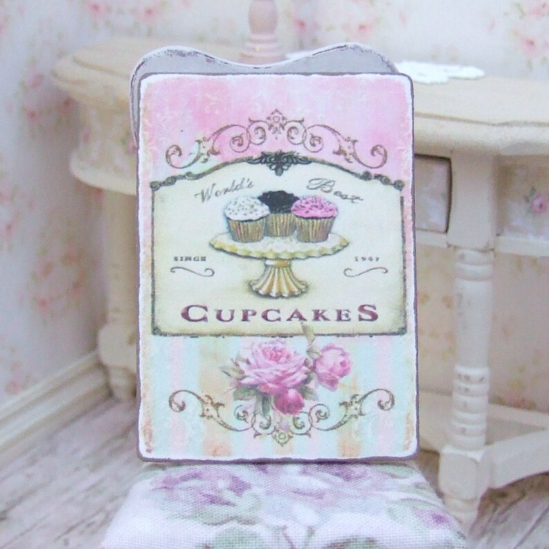 K&ouml;nnte beinhalten: Ein rechteckiges Schild mit rosa und hellblauem Hintergrund, mit dem Text "World's Best Cupcakes" und einer Illustration von drei Cupcakes auf einem Podest. Das Schild enth&auml;lt auch ein Blumendesign und die Worte "Since 1947".