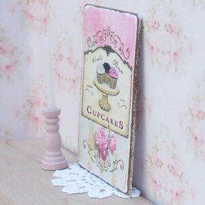 K&ouml;nnte beinhalten: Rechteckiges Holzschild mit rosa-cremefarbenem Streifenhintergrund, das ein Cupcake-Design und die Worte "World's Best Cupcakes" zeigt. Das Schild hat einen Used-Look mit goldfarbenem Rand und floralem Design.