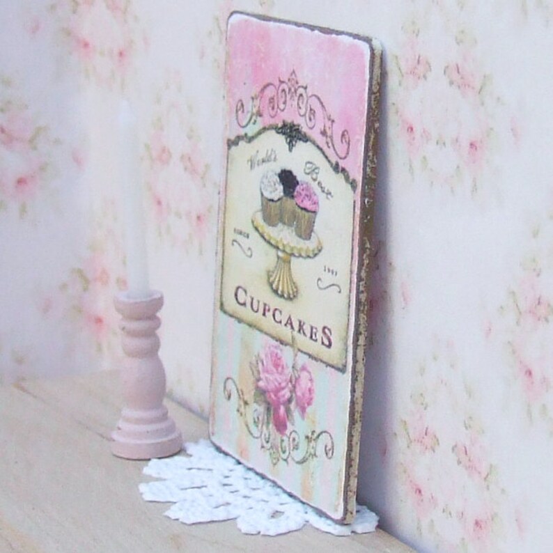 K&ouml;nnte beinhalten: Rechteckiges Holzschild mit rosa-cremefarbenem Streifenhintergrund, das ein Cupcake-Design und die Worte "World's Best Cupcakes" zeigt. Das Schild hat einen Used-Look mit goldfarbenem Rand und floralem Design.