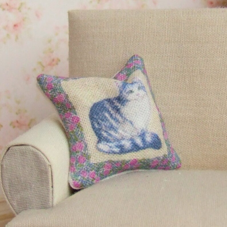 Op de afbeelding: Een klein, decoratief kussen met een blauw en wit kattenontwerp op een beige achtergrond. Het kussen heeft een roze en groene bloemenrand.