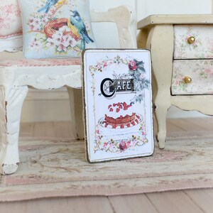 May include: A vintage-style sign with a brown wooden frame and a white background. The sign features a stylized illustration of a cake with strawberries and whipped cream, and the word "Cafe" in a decorative font.