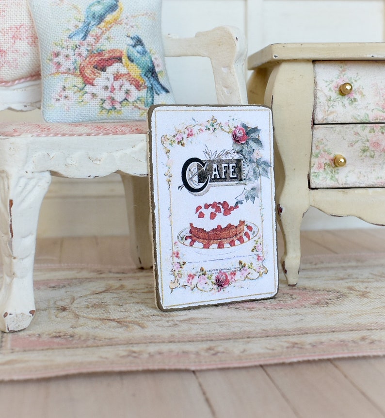 May include: A vintage-style sign with a brown wooden frame and a white background. The sign features a stylized illustration of a cake with strawberries and whipped cream, and the word "Cafe" in a decorative font.