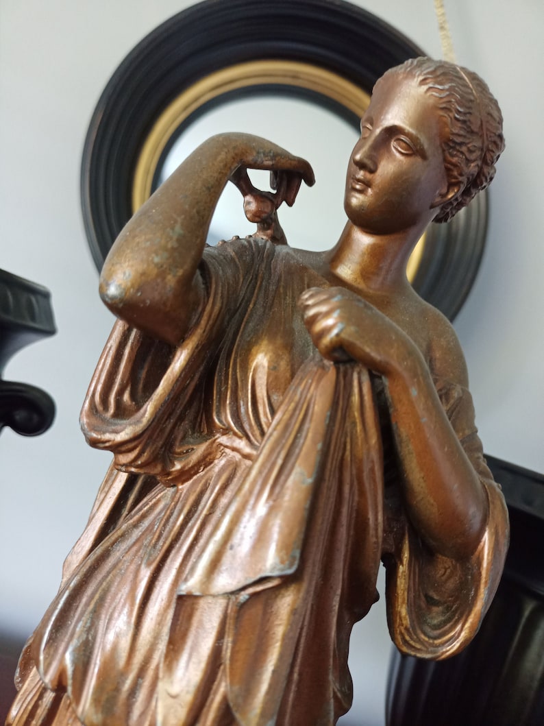 May include: A bronze statue of a woman in a flowing robe, holding a drapery in her hand. The statue has a detailed, realistic design and a warm, golden hue.