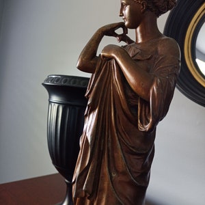 May include: A bronze statue of a woman in a flowing robe, standing with her hand on her chin, looking to the side. The statue is on a wooden surface next to a black vase.