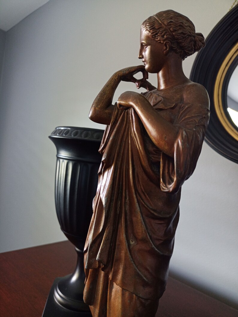 May include: A bronze statue of a woman in a flowing robe, standing with her hand on her chin, looking to the side. The statue is on a wooden surface next to a black vase.