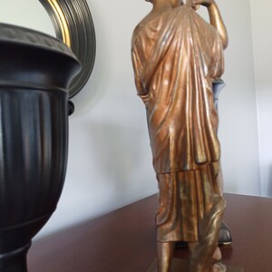 May include: A bronze-colored statue of a woman standing on a wooden base. The statue is wearing a long, flowing robe and has her hair pulled back in a bun. The statue is in a room with a black and gold mirror and a black vase.