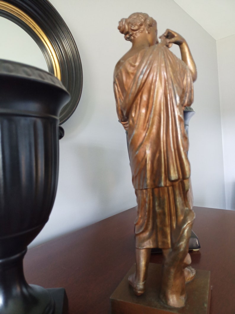 May include: A bronze-colored statue of a woman standing on a wooden base. The statue is wearing a long, flowing robe and has her hair pulled back in a bun. The statue is in a room with a black and gold mirror and a black vase.