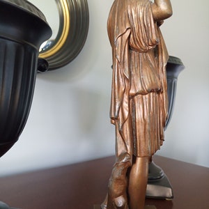 May include: A bronze statue of a woman in a flowing robe, standing on a base with a smaller bronze statue of a dog at her feet.