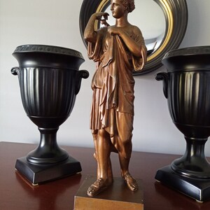 May include: A bronze statue of a woman in a flowing robe, standing on a wooden base. The statue is on a table with two black urns on either side.