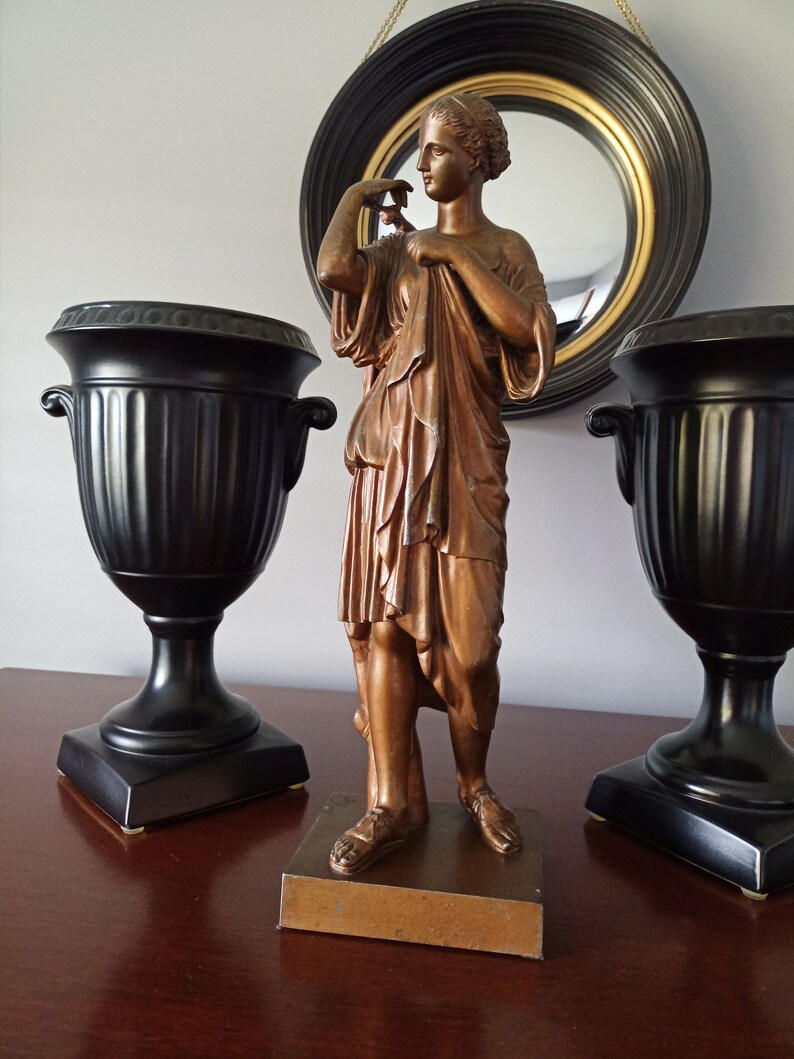 May include: A bronze statue of a woman in a flowing robe, standing on a wooden base. The statue is on a table with two black urns on either side.