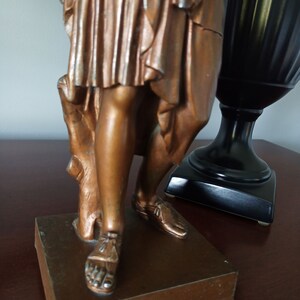 May include: A bronze statue of a woman standing on a square base. The woman is wearing a long flowing robe and sandals. The statue is in a vintage style.