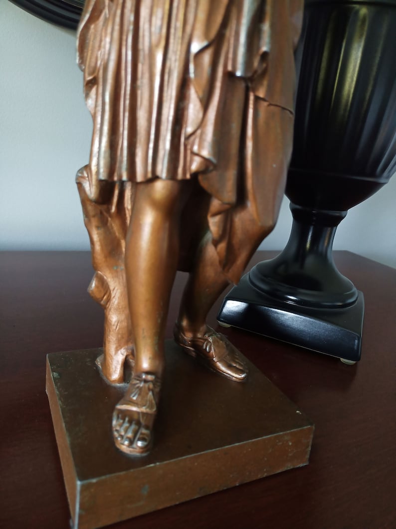 May include: A bronze statue of a woman standing on a square base. The woman is wearing a long flowing robe and sandals. The statue is in a vintage style.