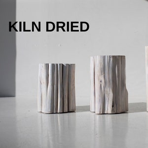 May include: Three cylindrical wooden stools with a weathered, light gray finish. The text "KILN DRIED" is printed in black above the stools. The stools vary in height and have a natural, textured appearance, suitable for home decor.