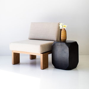 May include: A modern beige upholstered chair with a wooden frame. Beside it is a dark, geometric side table. A small, patterned vase with white flowers sits on the table, creating a minimalist interior design.