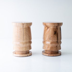 May include: Two wooden stools with a light brown finish. The stools have a cylindrical shape with a slightly wider top and base. The wood grain is visible, adding to the natural aesthetic. The stools are suitable for indoor use.