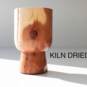 May include: A handcrafted wooden stool with a cylindrical top and a flared base. The stool is made of light brown wood with visible grain and knots. The text "KILN DRIED" is visible on the right side of the stool.