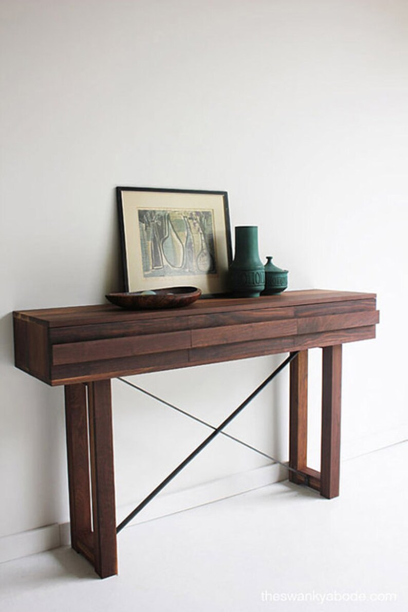 May include: A dark wood console table with two drawers and a black metal X-shaped base. The table is decorated with a framed print, a green vase, and a green ceramic jar.