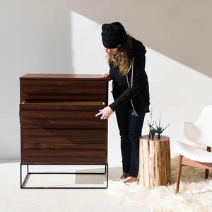 Pu&ograve; includere: Una cassettiera in legno scuro con quattro cassetti e una struttura in metallo nero. Il cassetto superiore &egrave; aperto, rivelando un interno di colore chiaro. Una sedia bianca con gambe in legno e un tavolino in legno sono vicini.