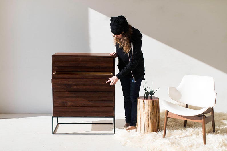 Pu&ograve; includere: Una cassettiera in legno scuro con quattro cassetti e una struttura in metallo nero. Il cassetto superiore &egrave; aperto, rivelando un interno di colore chiaro. Una sedia bianca con gambe in legno e un tavolino in legno sono vicini.